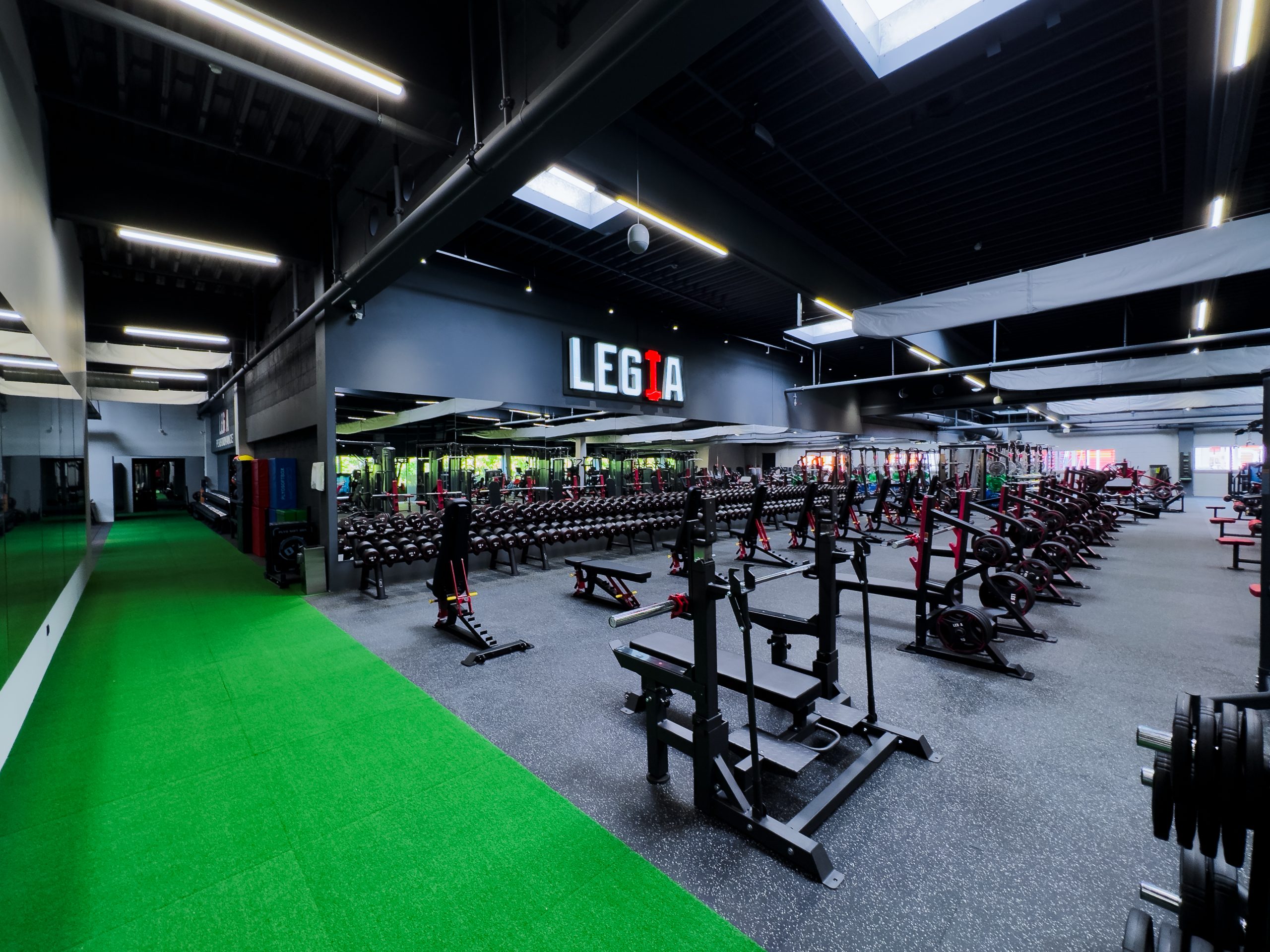 Interiér fitness centra Legia Fitness s pohľadom na profesionálnu silovú zónu, činkáreň a zelenú dráhu s umelou trávou pre funkčný tréning.